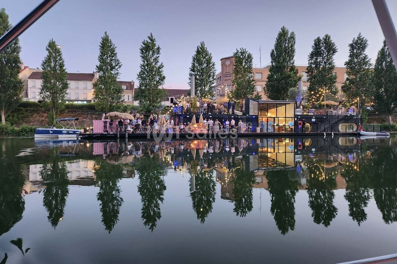 Des personnes rassemblées sur une péniche illuminée au bord d'un canal, entourée d'arbres et de bâtiments.