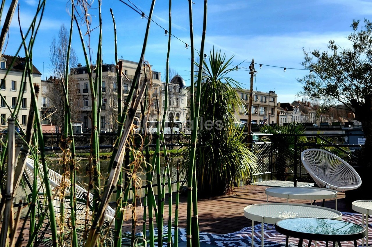 Terrasse aménagée avec mobilier extérieur, plantes et vue sur des bâtiments anciens près d'un canal sous un ciel bleu.