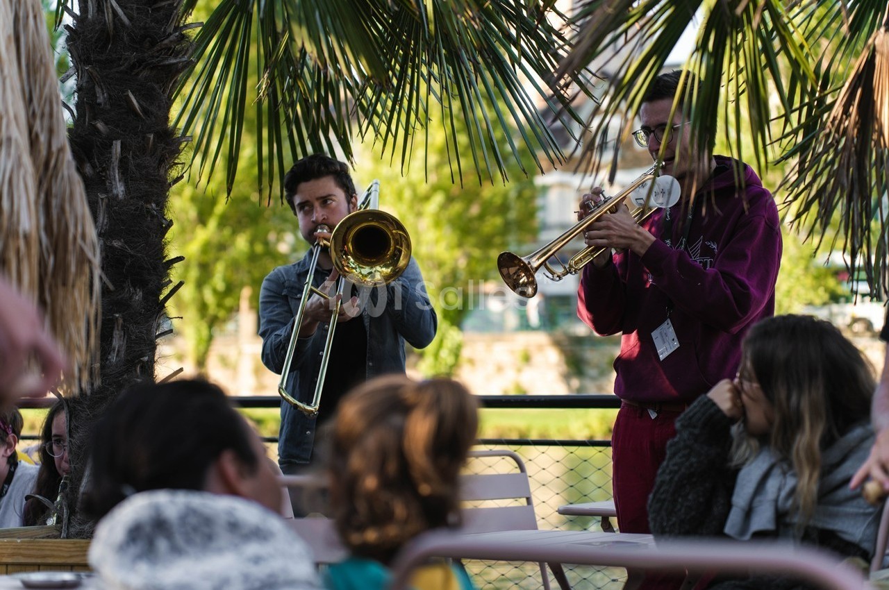 Deux musiciens jouent du trombone et de la trompette en plein air devant un petit public sous des palmiers.