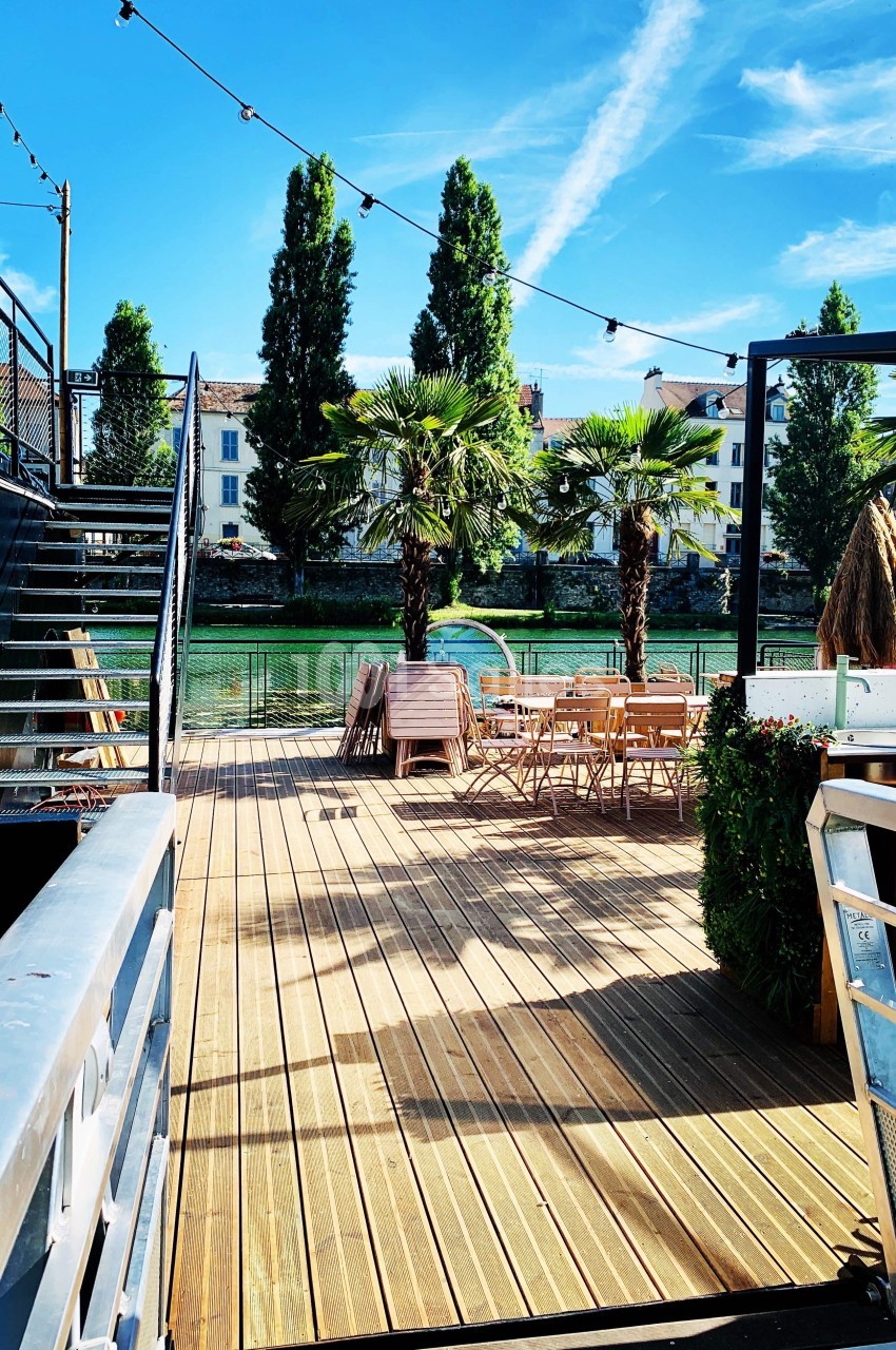 Terrasse en bois avec tables et chaises, bordée de palmiers, donnant sur un canal sous un ciel bleu.