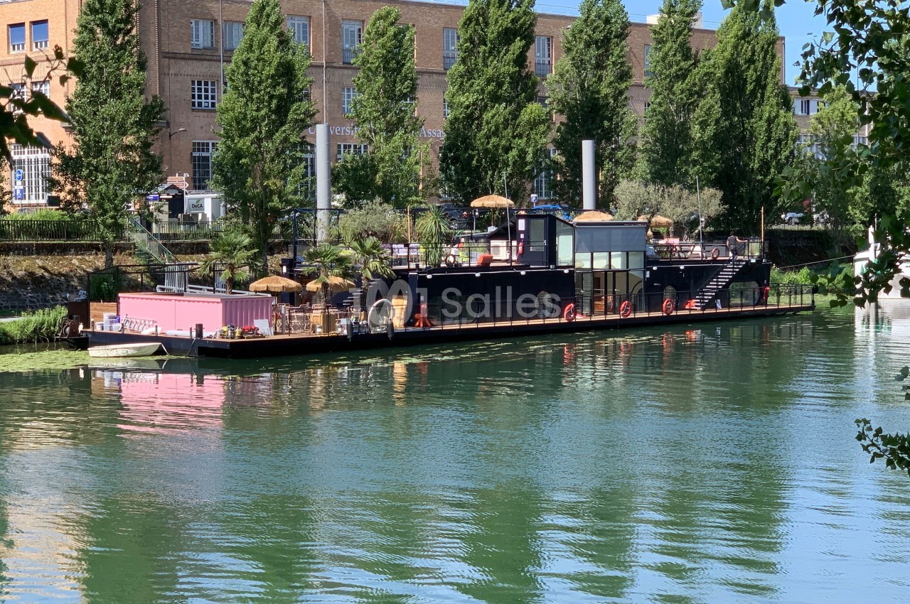 Bateau aménagé en terrasse flottante avec parasols, amarré sur une rivière bordée d'arbres et de bâtiments en arrière-plan.