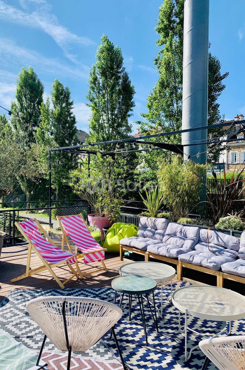 Terrasse ensoleillée avec transats colorés, canapé à coussins, tables basses et végétation environnante.