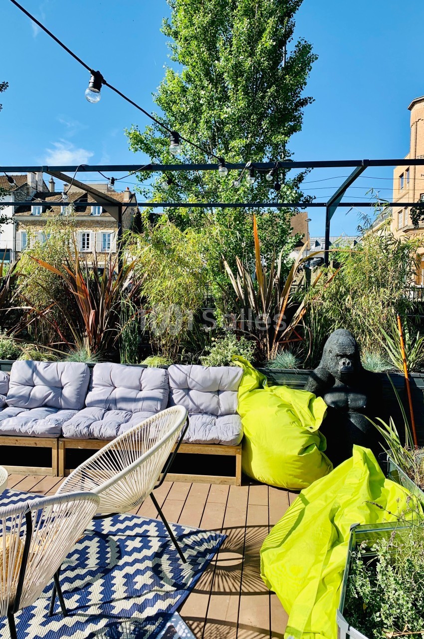 Terrasse aménagée avec canapé, fauteuils, coussins, plantes, gorille décoratif et poufs verts sous un ciel dégagé.