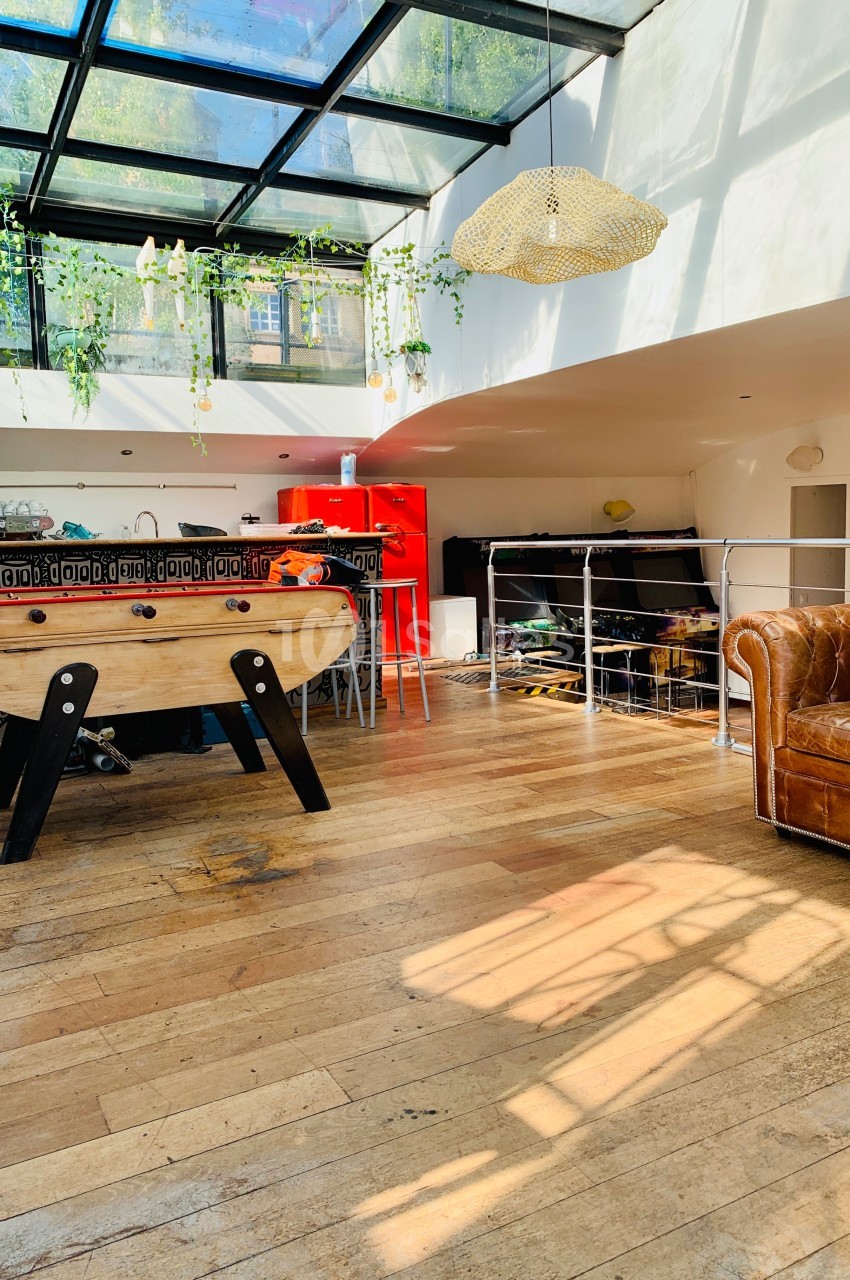 Espace lumineux avec parquet, baby-foot, canapé en cuir, cuisine rouge et verrière laissant entrer la lumière naturelle.