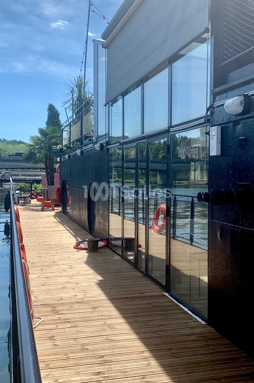 Vue latérale d'une péniche avec terrasse en bois, baies vitrées et bouées de sauvetage, amarrée au bord d'une rivière.