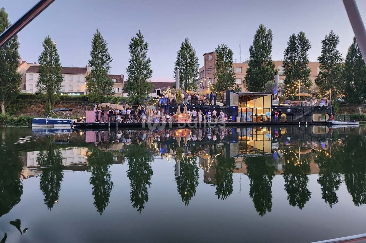 Personnes rassemblées sur une péniche éclairée au crépuscule, amarrée près d'arbres et de bâtiments au bord de l'eau.