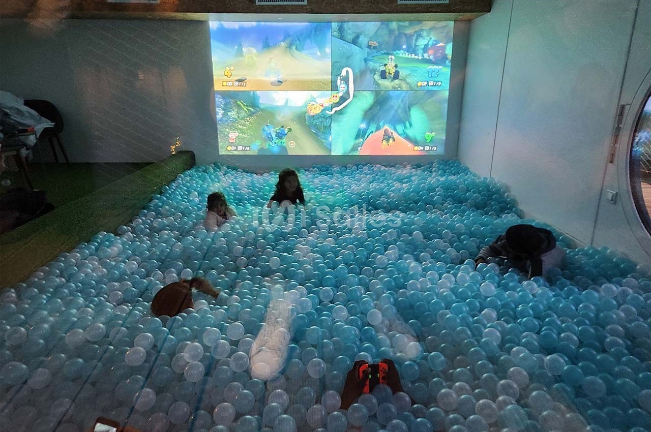 Des enfants jouent dans une piscine à balles bleues devant un écran projetant un jeu vidéo multijoueur.