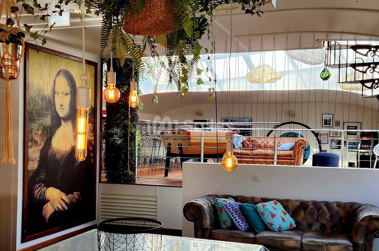 Salon lumineux avec canapé en cuir, plantes suspendues, tableau de la Joconde et mezzanine aménagée en arrière-plan.