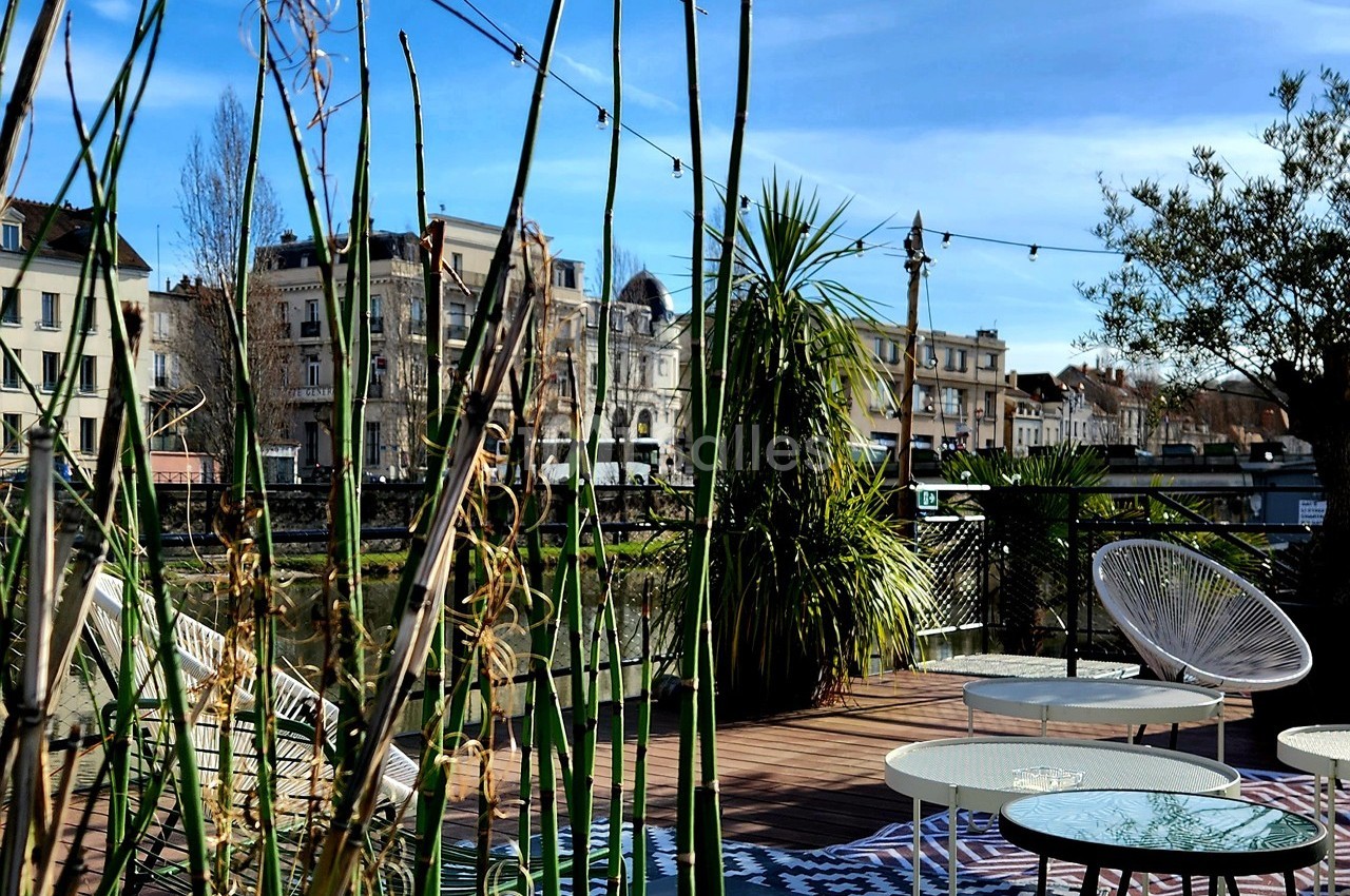 Terrasse aménagée avec des plantes, des tables et des chaises, vue sur des bâtiments et un cours d'eau en arrière-plan.