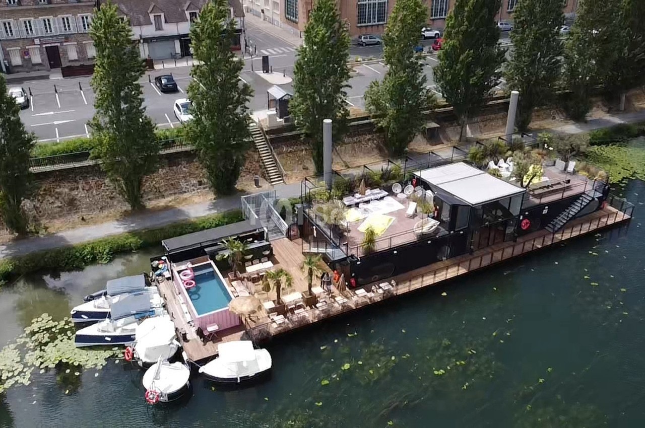 Vue aérienne d'une péniche aménagée avec terrasse, piscine et espaces de détente, amarrée près d'une rive arborée.