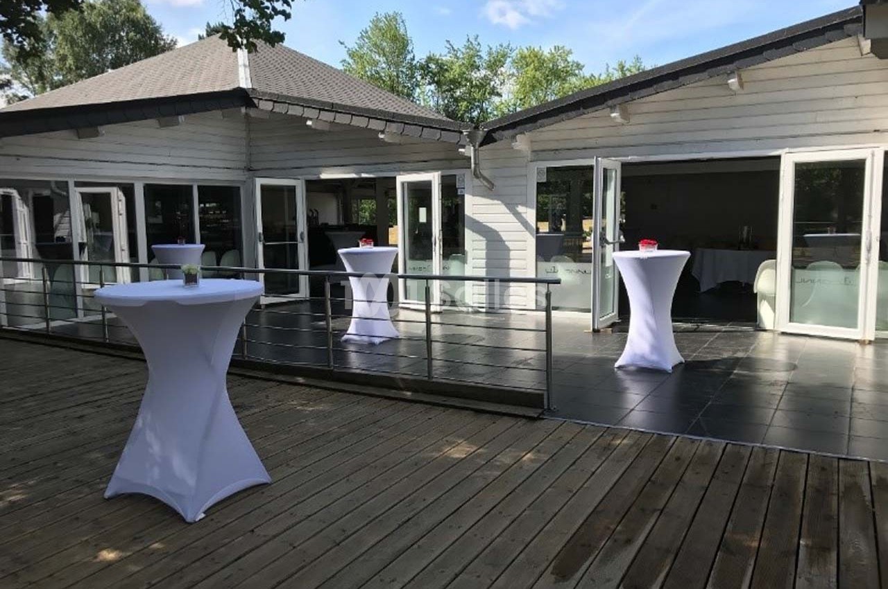 Terrasse en bois avec tables hautes couvertes de nappes blanches, devant un bâtiment moderne avec baies vitrées.