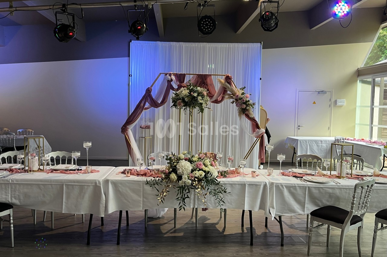 Salle décorée pour un événement avec tables dressées, arche florale et éclairage d'ambiance.