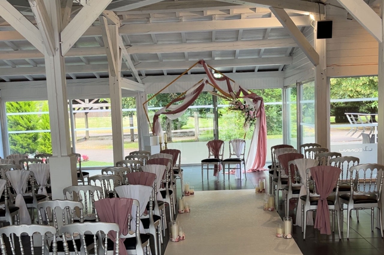 Salle de cérémonie décorée avec des chaises blanches ornées de rubans roses et une arche florale devant des baies vitrées.