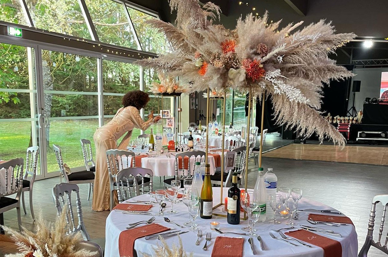 Salle de réception décorée avec des tables élégantes, des centres de table floraux et une femme ajustant un détail.