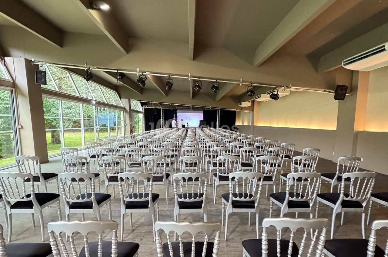 Salle de conférence lumineuse avec des rangées de chaises blanches alignées face à un écran de projection.