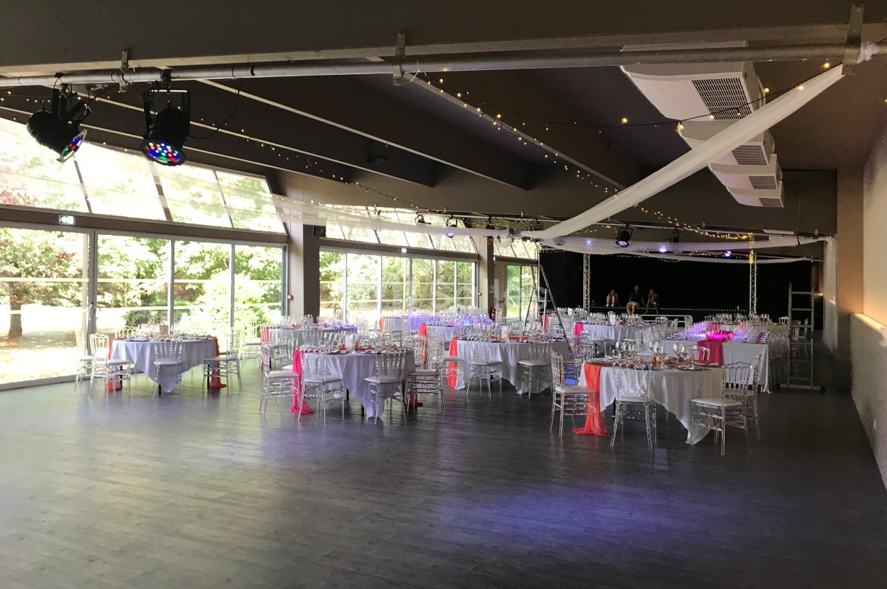 Salle de réception lumineuse avec grandes baies vitrées, tables dressées et décorations roses et blanches.