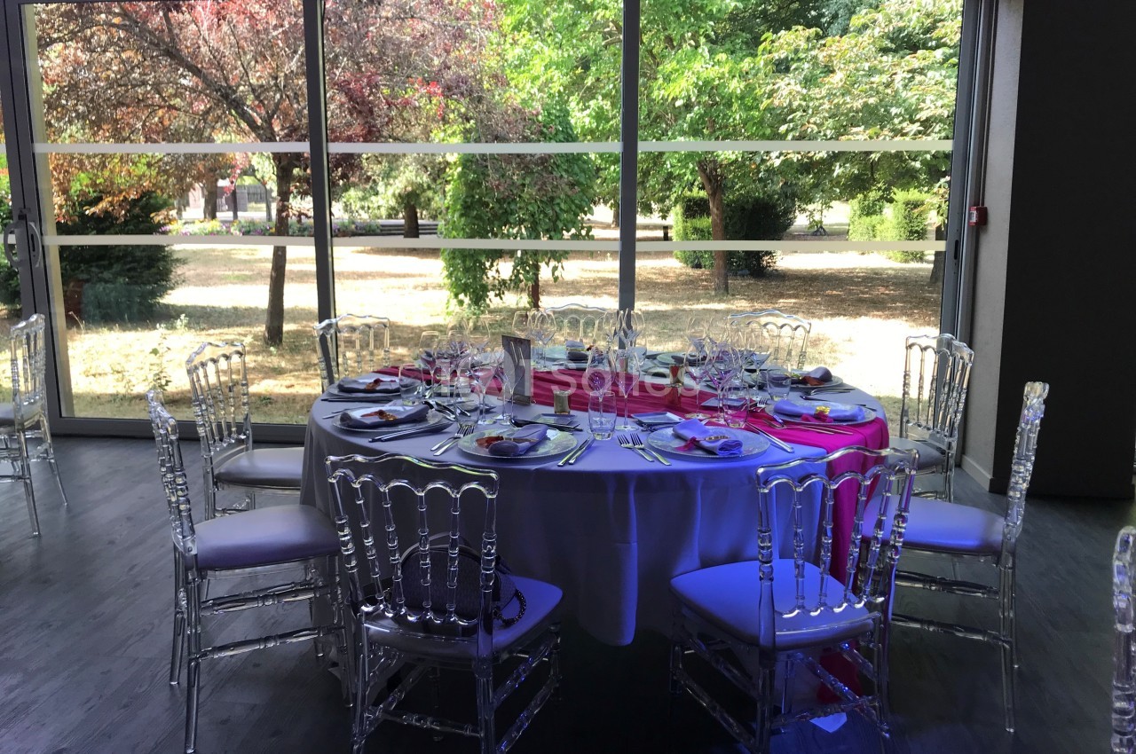 Table ronde dressée pour un repas, entourée de chaises transparentes, devant une baie vitrée donnant sur un jardin.