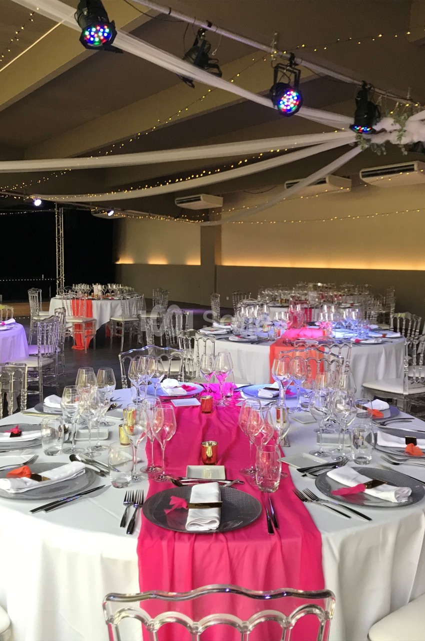 Salle de réception décorée avec des tables rondes, nappes blanches, chemins de table roses et chaises transparentes.