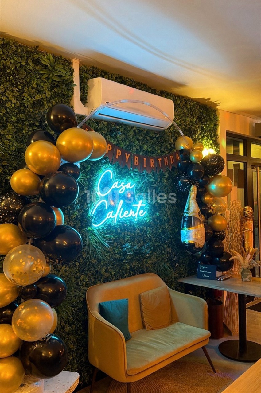 Mur végétal décoré avec des ballons noirs et dorés, néon ’Casa Caliente’, fauteuil et table dans un espace intérieur.