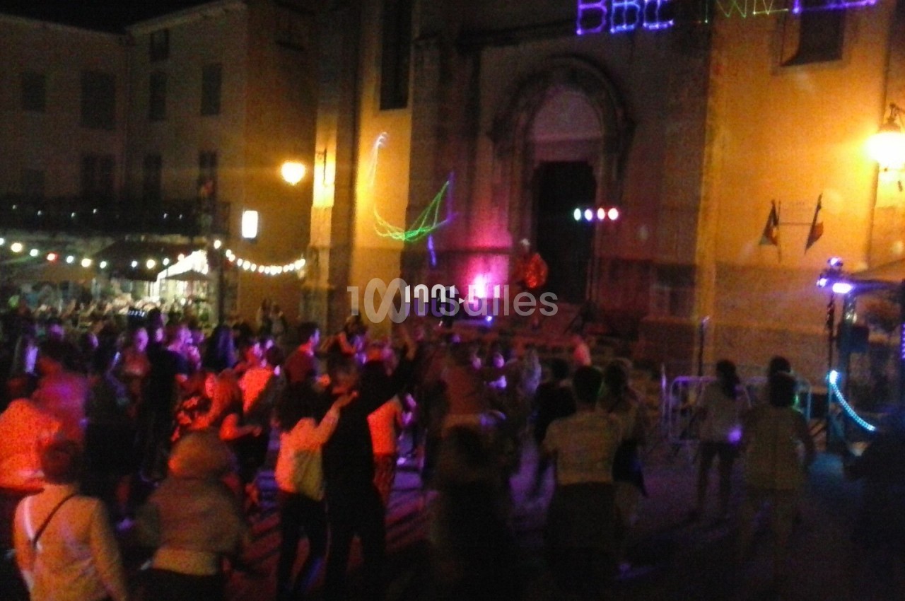 Foule dansant de nuit sur une place éclairée, devant une église avec des lumières colorées.