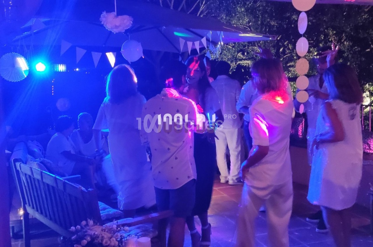 Des personnes en tenue blanche dansent et discutent sous des lumières colorées lors d'une soirée en extérieur.