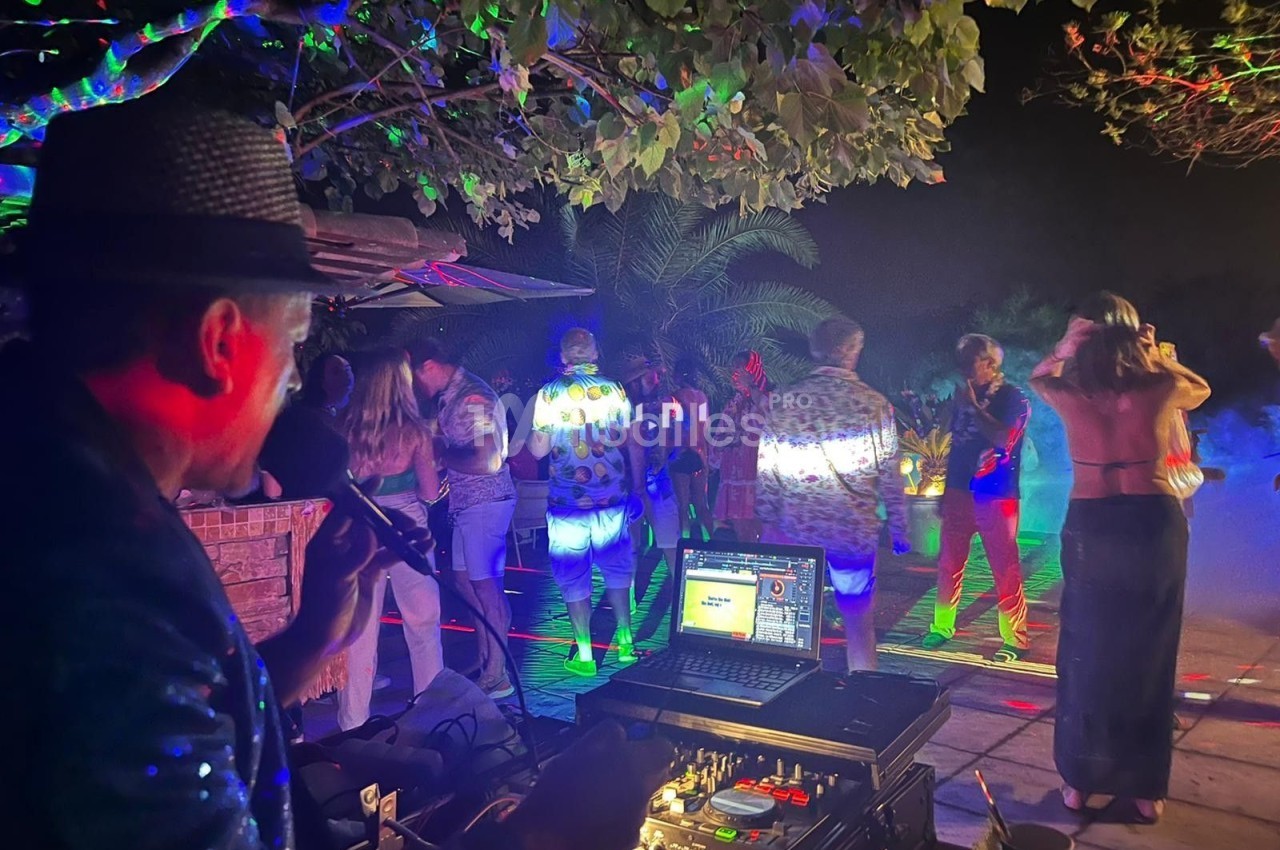 Un DJ anime une soirée en plein air avec des lumières colorées et des invités dansant sous des arbres.