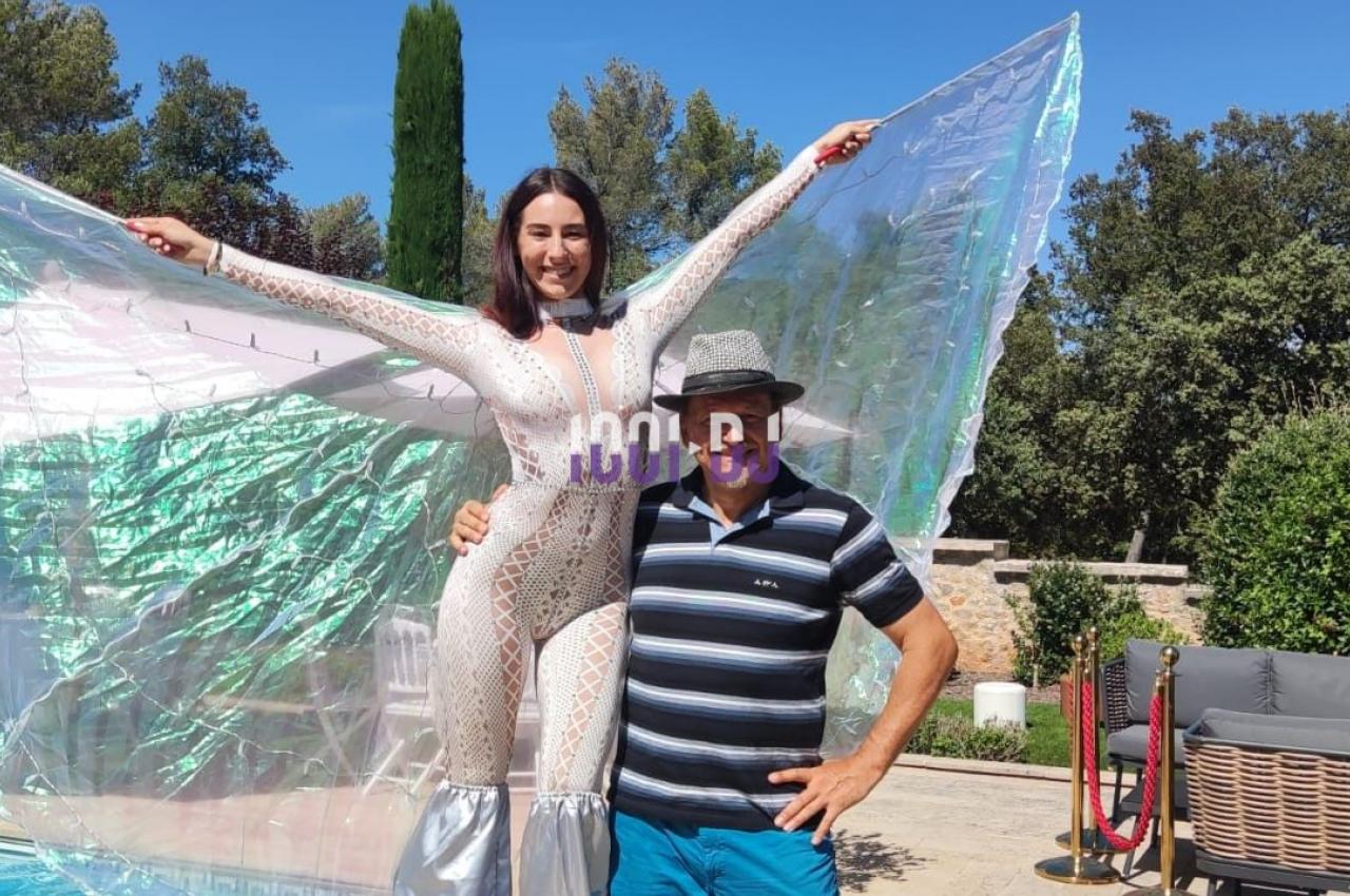 Une femme en costume scintillant pose sur des échasses à côté d'un homme, près d'une piscine en extérieur.
