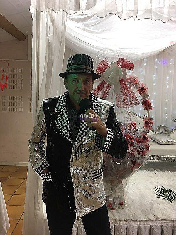 Homme en costume brillant noir et argent, tenant un téléphone devant un décor festif avec fleurs et voilage blanc.