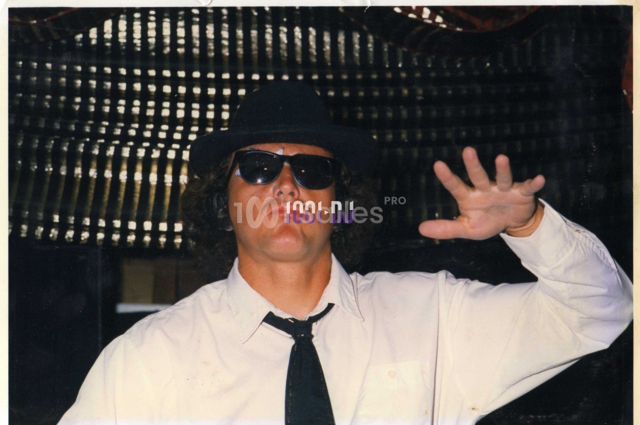 Homme portant des lunettes de soleil, un chapeau noir et une cravate, levant une main devant un fond sombre.
