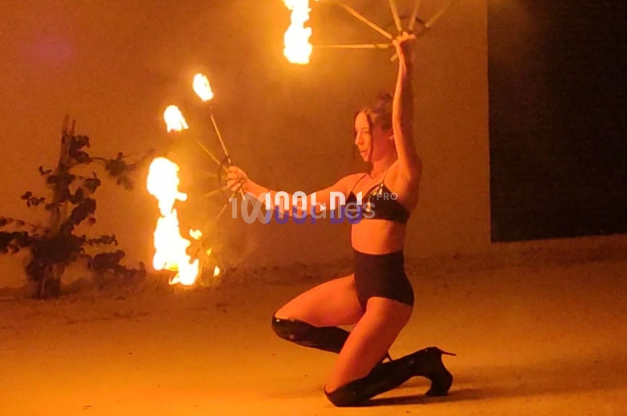 Une femme en tenue noire manipule des éventails enflammés, agenouillée dans un espace extérieur éclairé par les flammes.