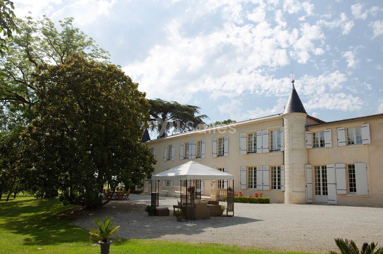 Façade d'un château en pierre avec tours, entouré d'arbres, terrasse aménagée avec parasol au premier plan.
