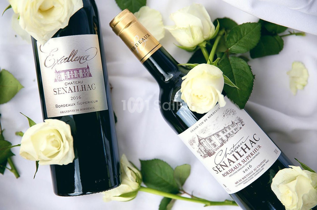 Deux bouteilles de vin rouge Château Senailhac entourées de roses blanches sur un fond clair.