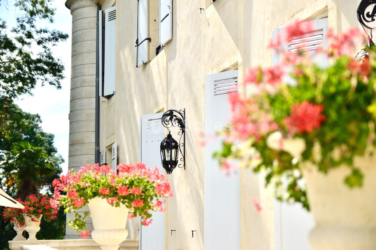 Façade ensoleillée d'un bâtiment avec volets blancs, lanternes murales et jardinières fleuries au premier plan.