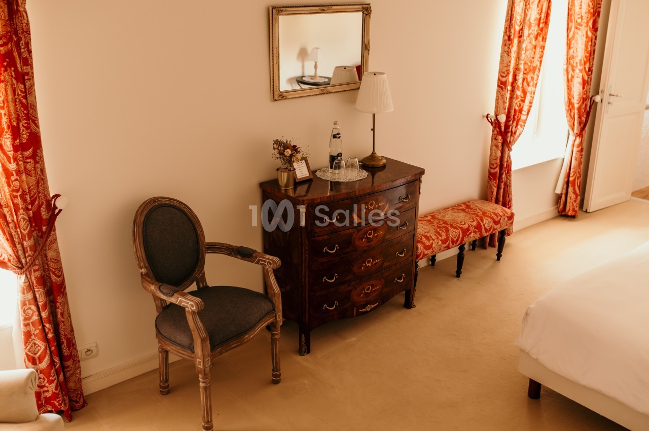 Chambre lumineuse avec commode en bois, chaise, miroir, rideaux rouges et lit partiellement visible.