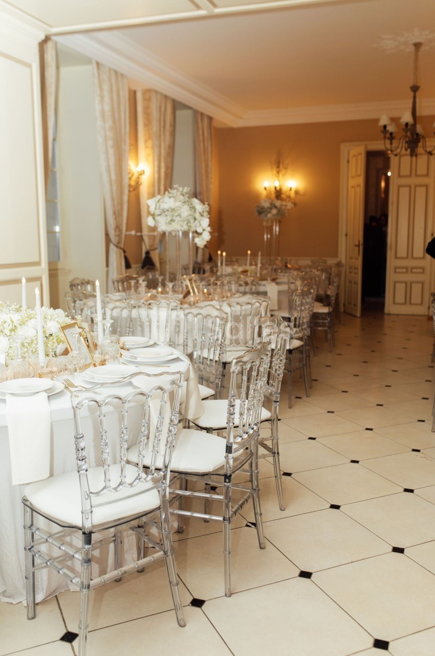 Salle élégante avec tables rondes dressées, chaises transparentes et décoration florale blanche dans un cadre lumineux.