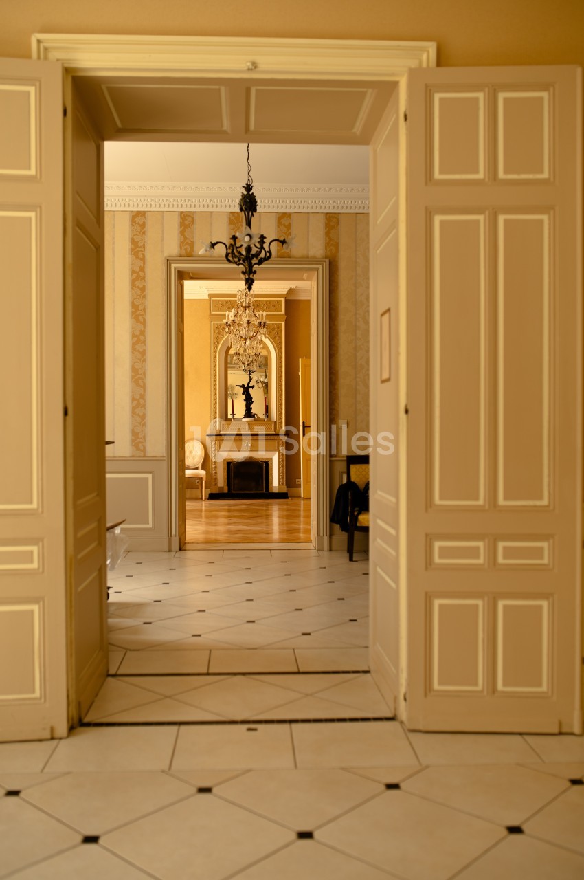Vue intérieure d'une pièce avec des portes ouvertes, révélant un salon lumineux avec cheminée et lustre.
