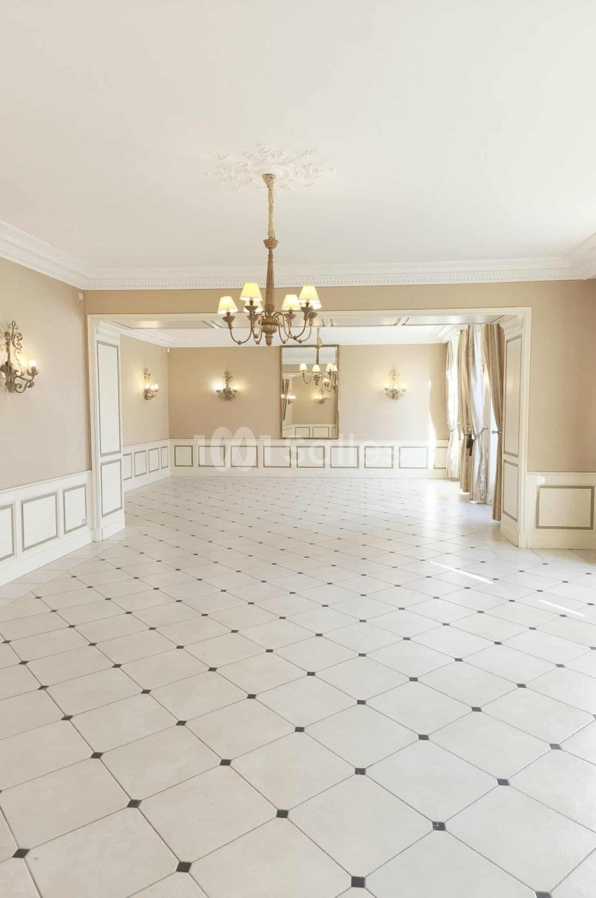 Salle lumineuse avec carrelage clair, moulures au plafond, lustres et grandes fenêtres avec rideaux beiges.