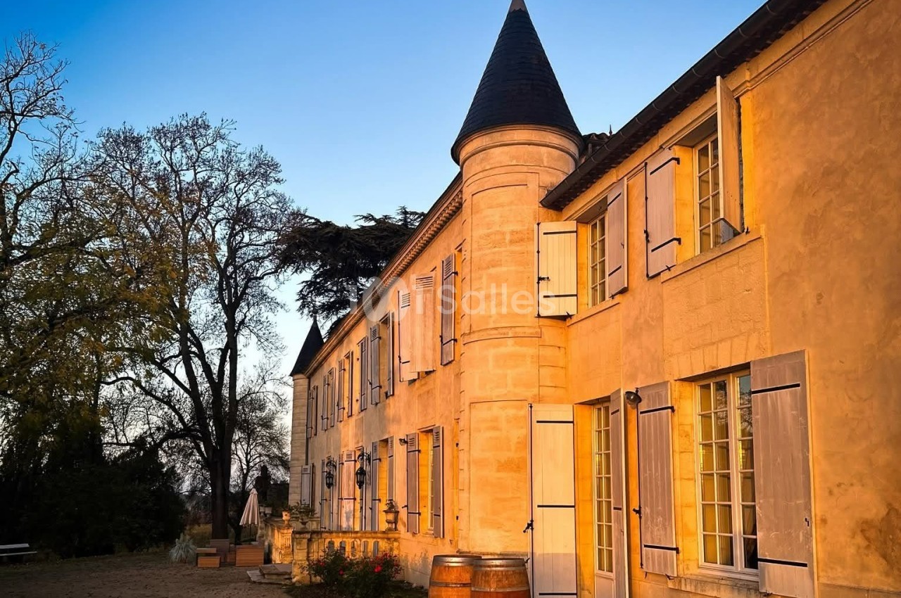 Façade d'un bâtiment en pierre avec des volets ouverts, éclairée par une lumière chaude au coucher du soleil.