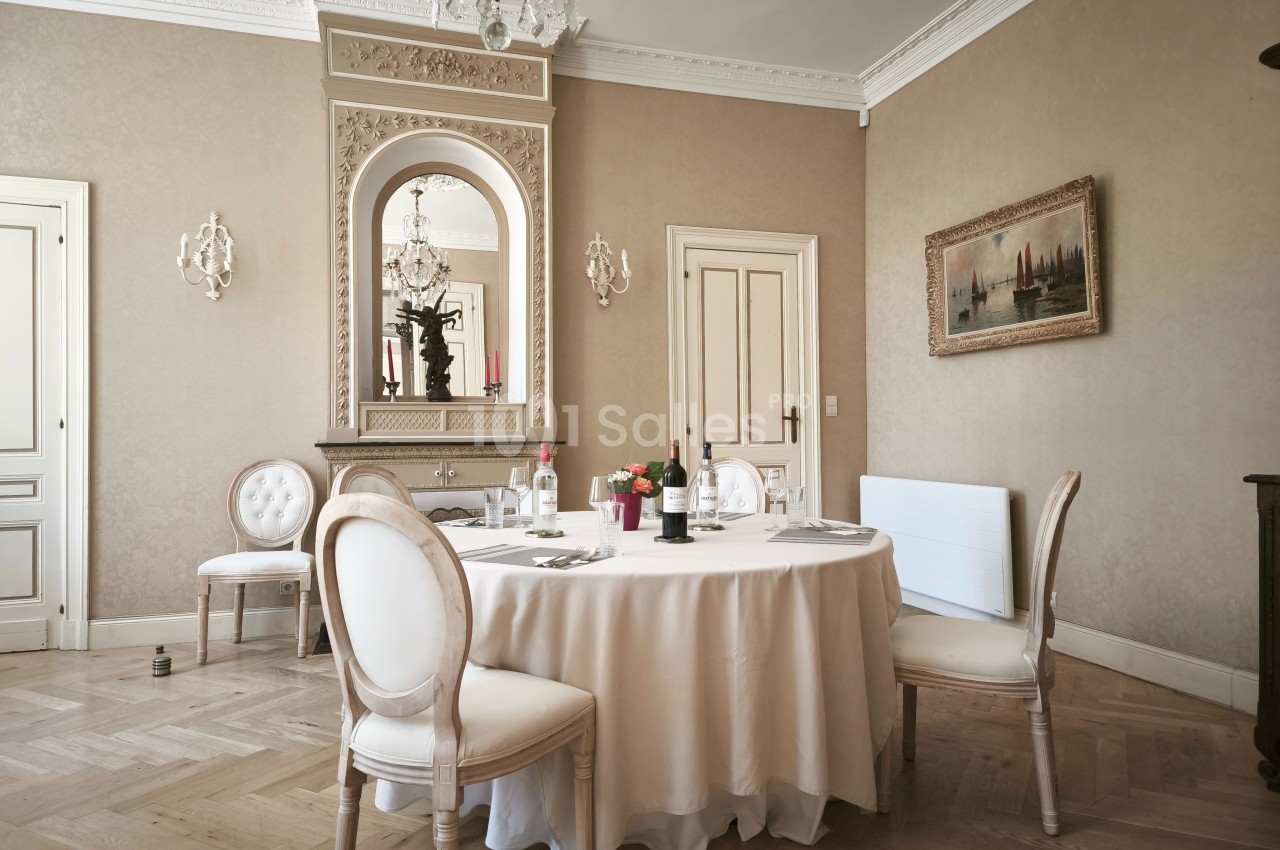 Salle à manger élégante avec table ronde dressée, chaises classiques, miroir orné et parquet clair.