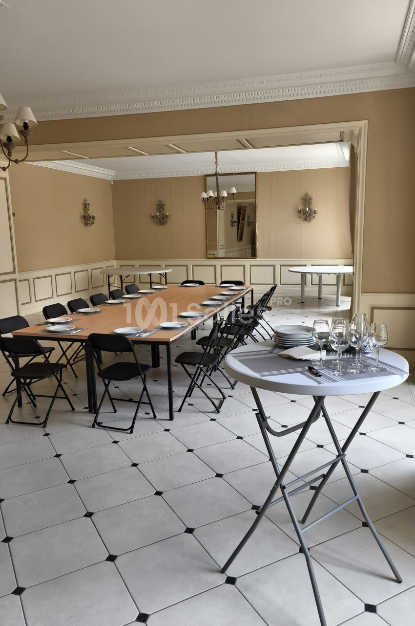 Salle de réunion lumineuse avec tables disposées en U, chaises pliantes noires et table ronde avec verres à vin.