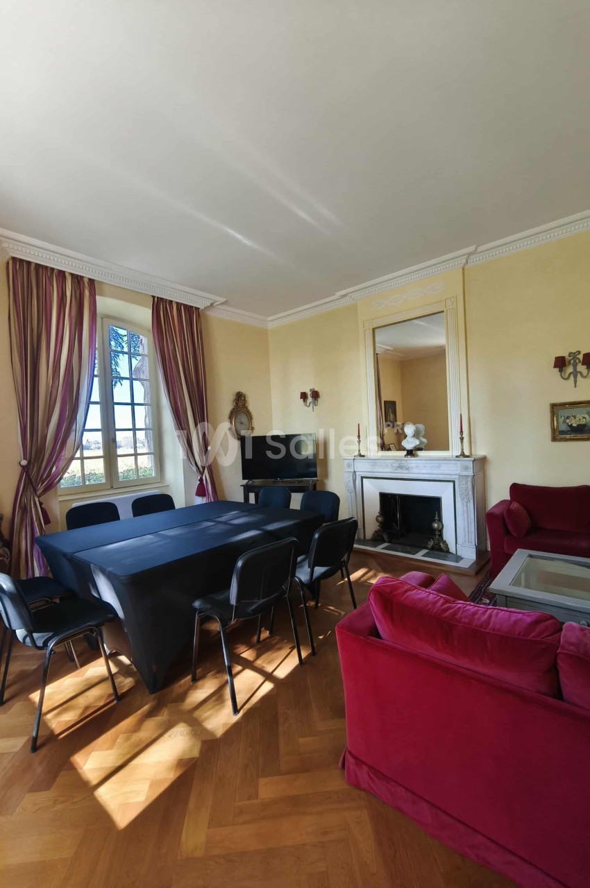 Salle lumineuse avec table rectangulaire, chaises noires, cheminée décorative, canapé rouge et grandes fenêtres.
