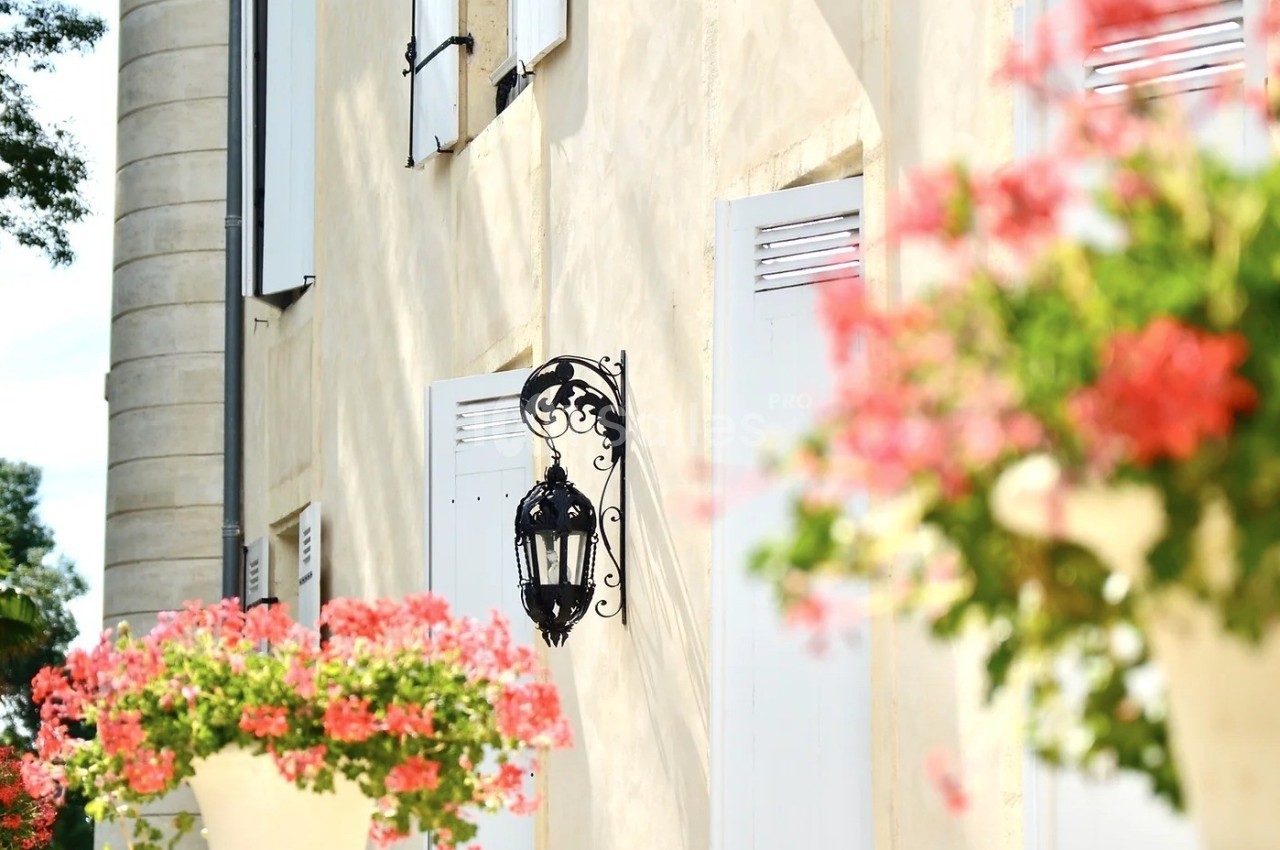 Façade claire avec volets blancs, lanterne en fer forgé et jardinières de fleurs roses en premier plan.