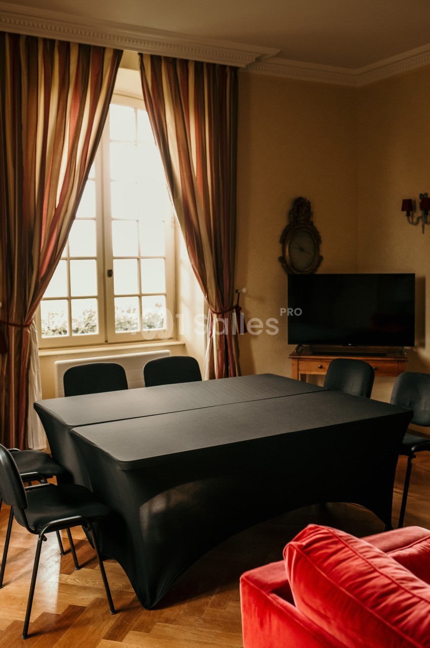 Salle avec une table noire recouverte, six chaises, un téléviseur, des rideaux rayés et une fenêtre lumineuse.