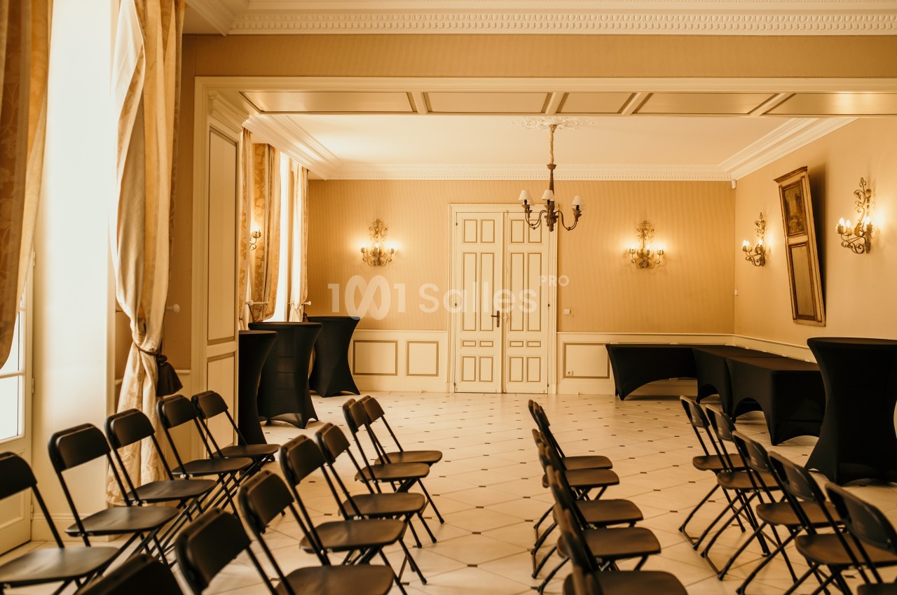 Salle lumineuse avec chaises noires alignées, tables couvertes de nappes noires et murs décorés de cadres et appliques.