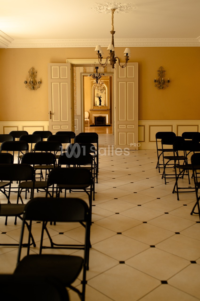 Salle lumineuse avec des chaises noires pliantes alignées, portes ouvertes sur une pièce avec cheminée et miroir.