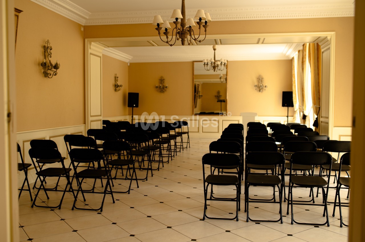 Salle de conférence avec rangées de chaises noires pliantes, éclairée par des lustres et la lumière naturelle.
