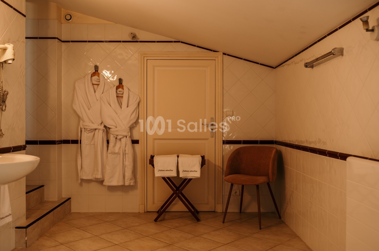 Deux peignoirs accrochés à une porte dans une salle de bain carrelée avec chaise et porte-serviettes.