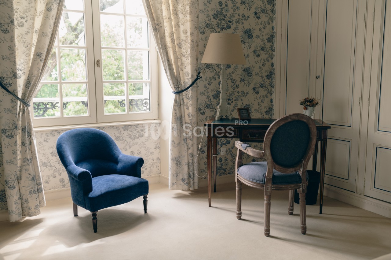 Fauteuil bleu, bureau en bois et chaise dans une pièce lumineuse avec papier peint fleuri et grandes fenêtres.