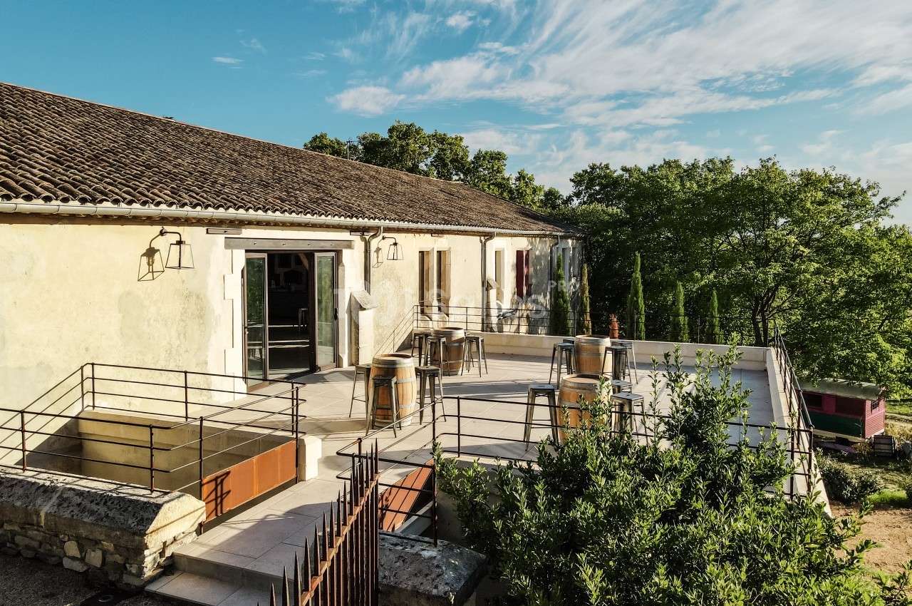 Terrasse ensoleillée avec tables hautes, chaises et vue sur des arbres, attenante à un bâtiment en pierre.