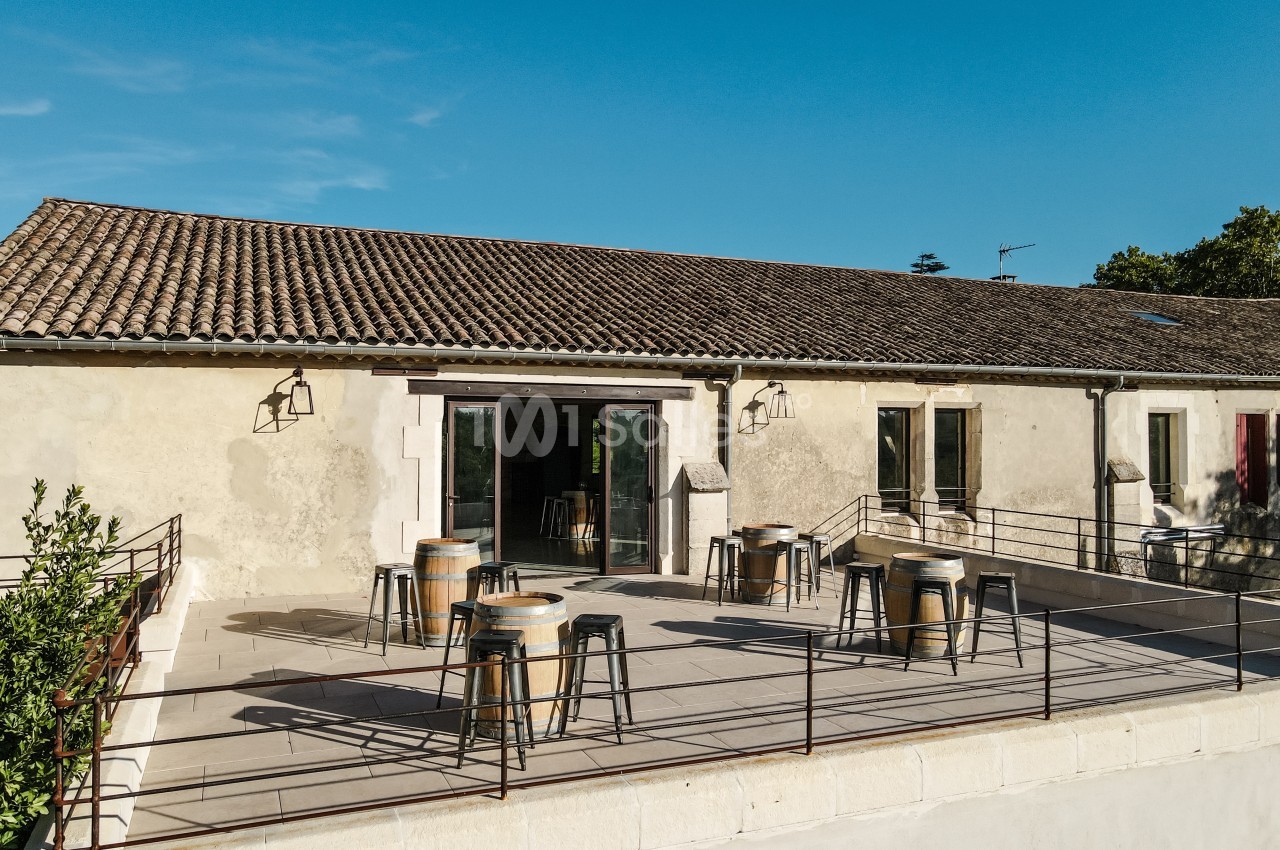Terrasse ensoleillée avec tables et tabourets, devant un bâtiment en pierre aux toits en tuiles.